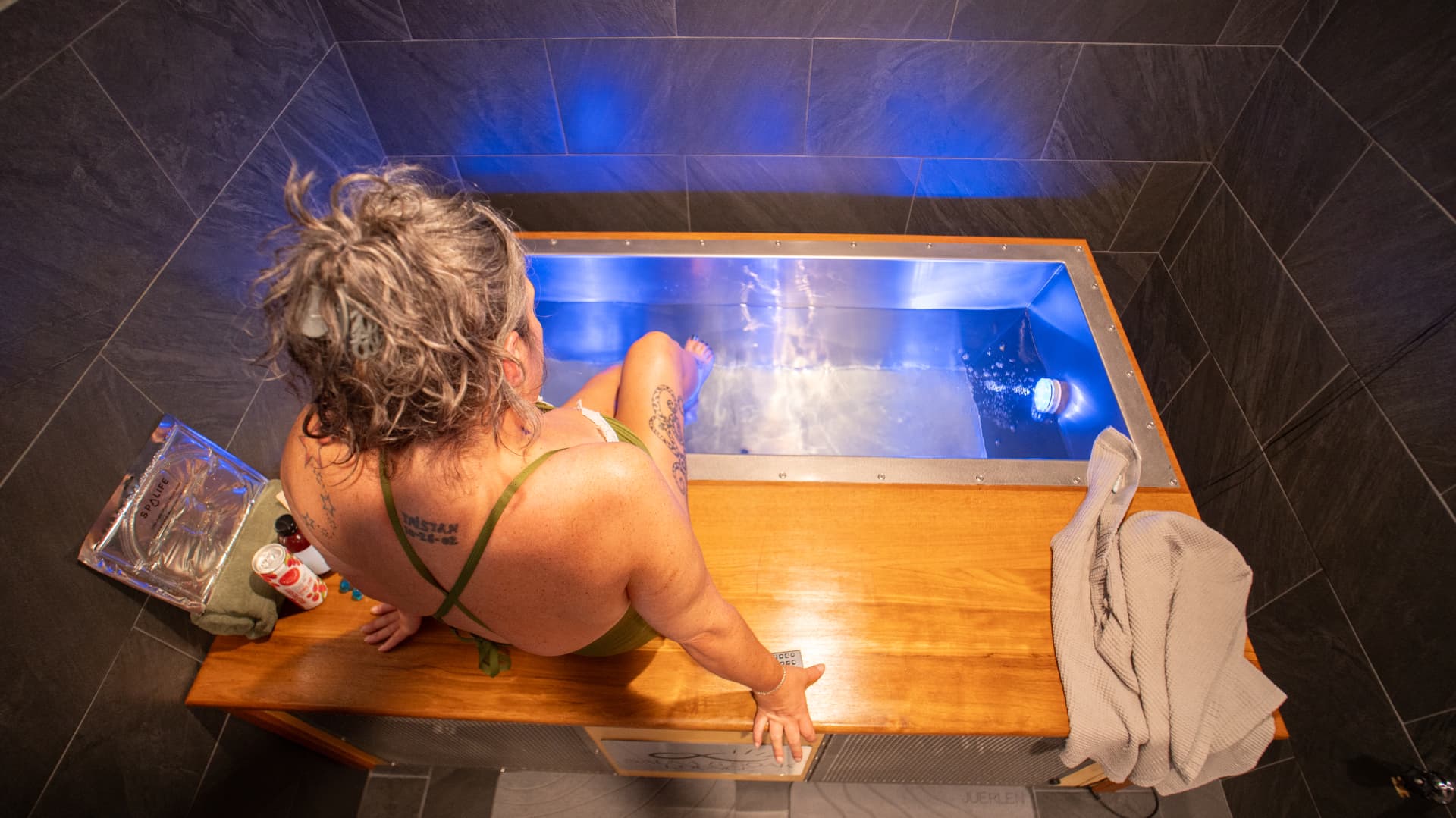 Overhead view of a guest seated on the timber bench beside the blue-lit cold plunge with towels and amenities