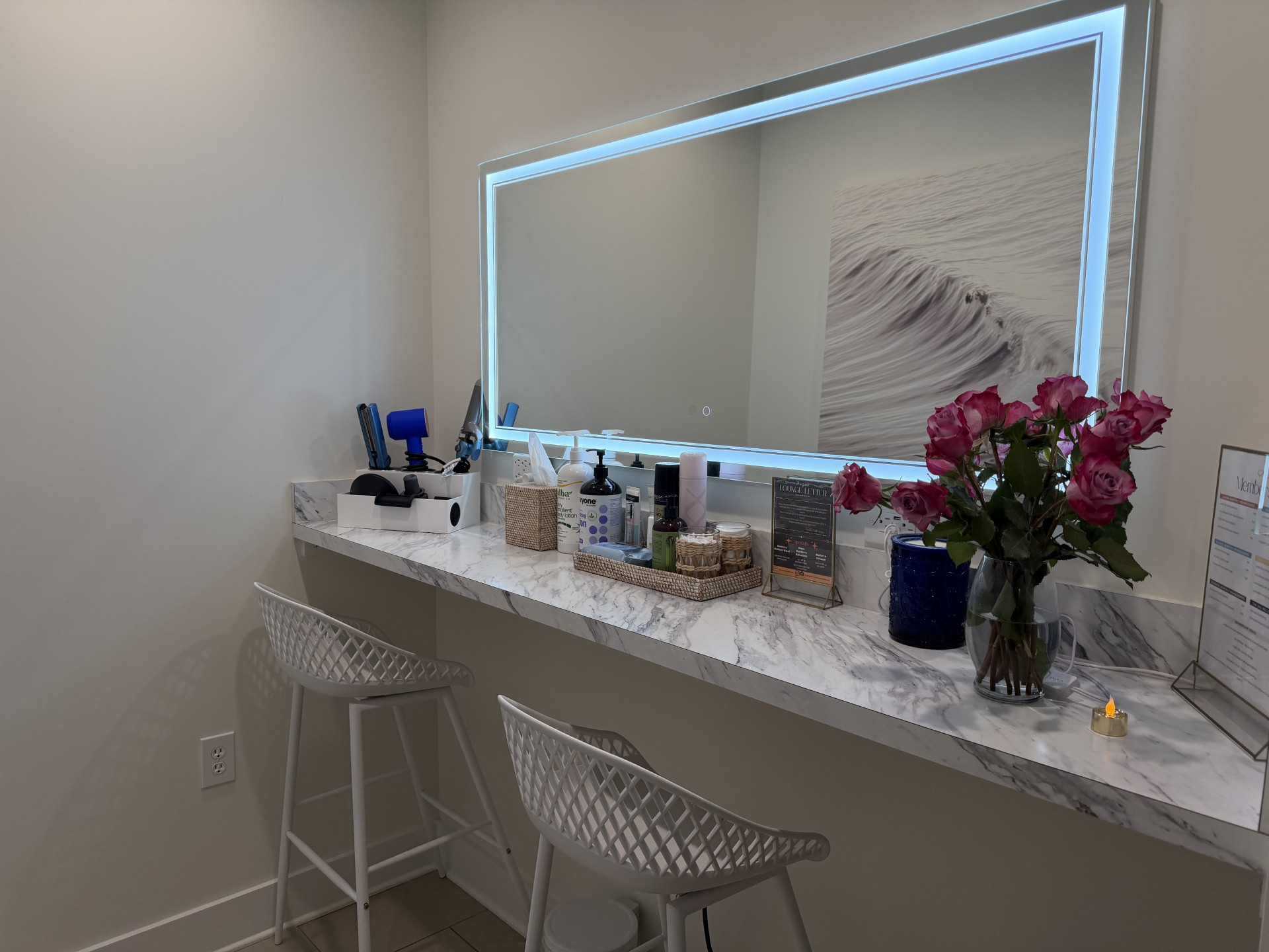 Marble vanity counter with an LED-lit mirror, styling tools, fresh roses, and white mesh chairs